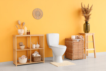 Interior of restroom with toilet bowl, shelving unit, basket and vase with pampas grass near orange wall