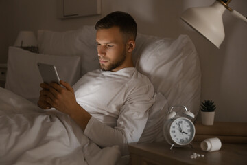 Young man suffering from insomnia and using tablet on bed in bedroom at late night