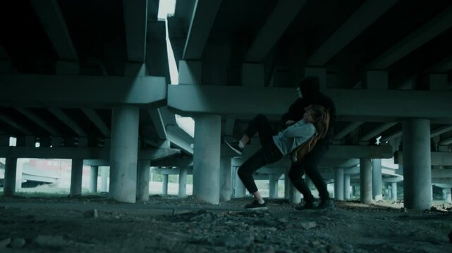 Side full shot of anonymous man in dark clothes and mask chasing and abducting young woman, sedating her with chloroform in desolated urban place under bridge