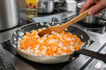 A close-up of a frying pan on a stove, filled with diced butternut squash and onions being sautéed. A wooden spoon stirs the colorful mixture in a well-equipped kitchen.