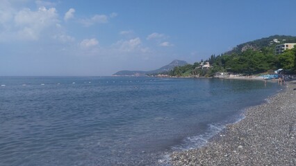 The wave, foaming, rolls onto the pebble beach of the seashore. The mountains in the bay are in a haze. People swim in the sea. There are pine trees and buildings on the shore. Sunny autumn weather
