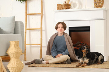 Little boy with warm blanket and cute cocker spaniel sitting near fireplace at home