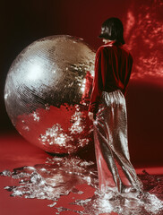A model poses confidently with her hands on an oversized disco ball, exuding vintage charm against a dark red backdrop.