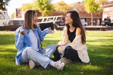 Two women friends sisters sitting in the park and have emotional talk conversation sitting in the grass. Best friends have emotional chat with gesticulating.