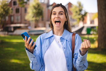 Blonde short hair woman browsing internet with cellphone mobile phone wearing casual clothes shirt gesturing like winner. Smiling open mouth woman look happy, celebrating win, triumph, success, wow.