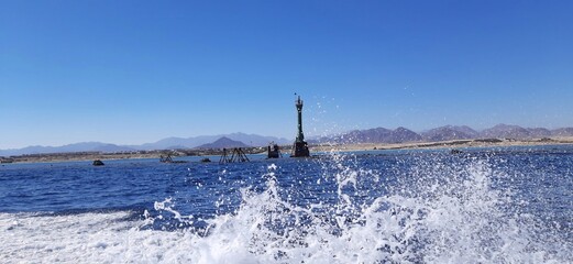 A serene view of a lighthouse near the coastline, surrounded by deep blue sea, with mountains in the background under a clear sky. Water splashes in the foreground.