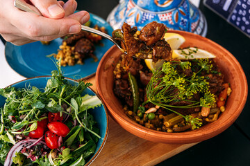 Traditional Moroccan lamb tagine served with a fresh salad. A hand scoops the dish, highlighting Mediterranean flavors and cultural culinary practices. © irissca