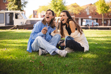 Two happy women friends sisters sitting in park on grass look at side point finger, laughing and show wow emotions. 