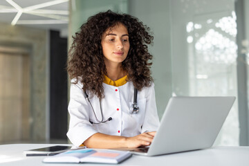 Young female doctor wearing white coat and stethoscope, working on laptop in modern office. Focused and professional atmosphere with documents and technology. Concept of healthcare innovation.