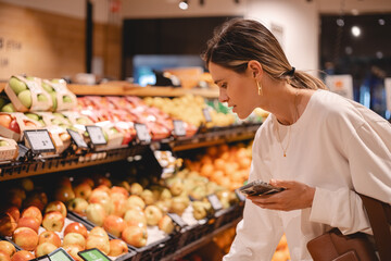 Young woman girl buyer client consumer stands in shop store supermarket choosing food buying puts in basket. Woman choose fruits.