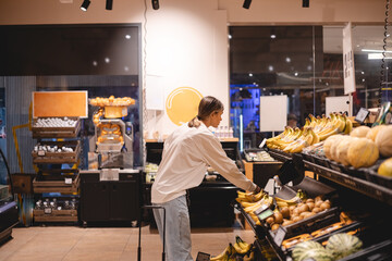 Young woman girl buyer client consumer stands in shop store supermarket choosing food buying puts in basket. Woman choose bananas.