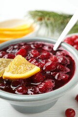 Tasty cranberry sauce in bowl and orange on white table, closeup