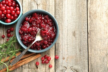 Tasty cranberry sauce in bowl, berries, rosemary and cinnamon sticks on wooden table, flat lay. Space for text