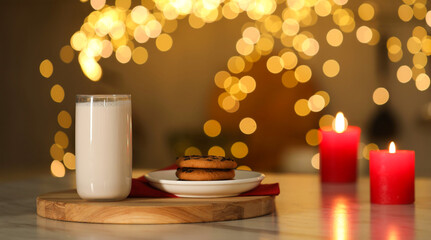Glass of milk, tasty chocolate chip cookies and burning candles on table against blurred Christmas lights. Bokeh effect