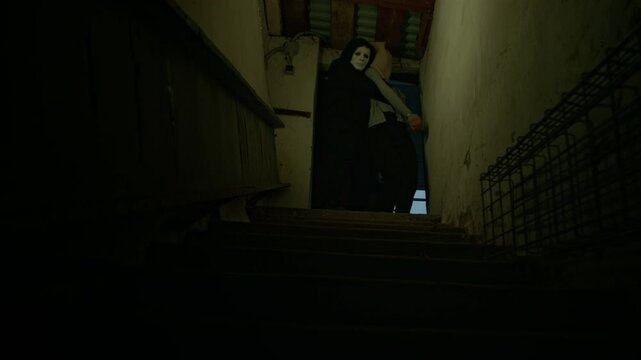 Full low-key shot of anonymous masked criminal carrying hostage with sack on head and tied hands, going down stairs into dark basement, human trafficking and abduction