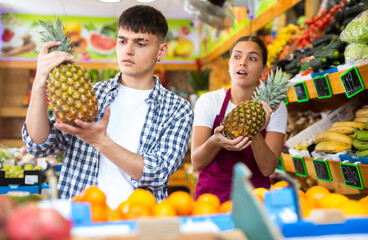 Young seller girl in apron selling fresh pineapple to man customer in grocery store