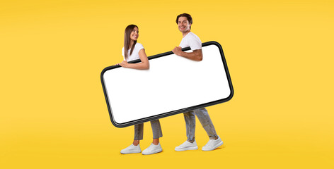 Cool Mobile App. Cheerful young couple holding big giant smartphone with mockup, walking together, promoting application or website, advertising your service or product over orange studio background
