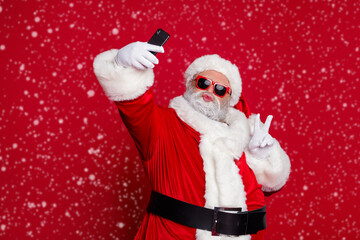 Photo of funky handsome santa claus wear red costume hat eyewear showing v-sign tacking selfie lips pouted isolated red color background