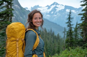 Naklejka premium A joyful woman with brown hair stands on a lush green hilltop, wearing a yellow backpack and grey jacket, overlooking a stunning snow-capped mountain range.
