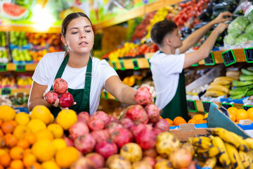 Happy young European woman in apron selling fresh pomegranate and fruits in hypermarket