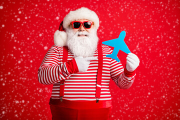 Photo of old man grey beard hold direct finger paper plane smile wear santa claus x-mas costume suspenders sunglass striped shirt cap isolated red color background