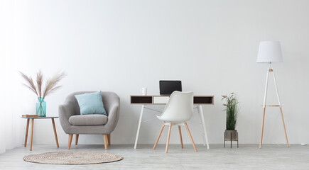 Home office, cabinet or workplace at house for remote work. Desk with laptop, white chair, simple armchair with blue pillow and table with vase and dry plants. Light walls in daylight, panorama