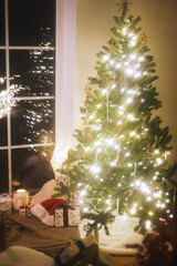 Merry Christmas! Stylish christmas tree with lights, glowing lantern, santa hat and stylish gifts under xmas tree in room. Atmospheric christmas eve at home, soft winter hygge still life