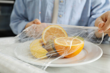 Woman putting plastic food wrap over plate with fruits at countertop in kitchen, closeup