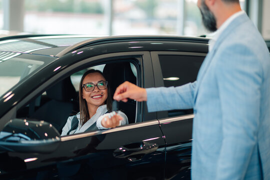 Woman in glasses smiling, receiving car keys from suited man in a dealership