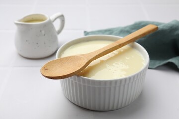 Tasty condensed milk in bowl and jug with spoon on white tiled table, closeup