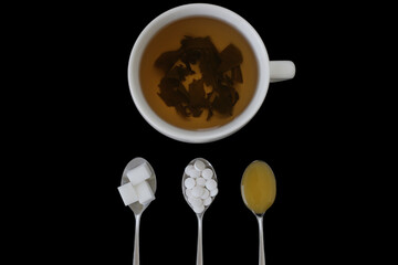 Cup of green tea and spoons with sugar, aspartame and honey on black background