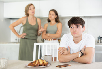 Young guy is sitting near kitchen table and talking to mother and sister arguing. Family conflict, turmoil, quarrel