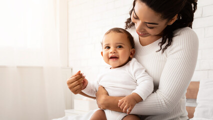 Joyful African American Mother Hugging Baby Playing With Charming Toddler Boy Posing Sitting On Bed In Bedroom Indoor. Child Care And Joy Of Being Mom Concept