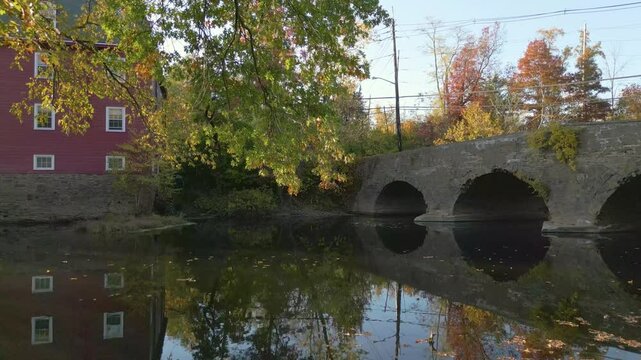Old stone bridge and historic mill on a tranquil river with Fall foliage during golden hour, drone 4K