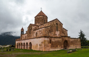 Historic Odzun church stands under a cloudy sky, surrounded by lush green grass and distant mountains, evoking a sense of serenity and timeless beautyю. Lori Region, Armenia