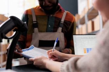 Storage room supervisor signing goods inventory report, african american coworker working at clients orders preparing packages in warehouse. Diverse team standing at counter in storehouse. Close up