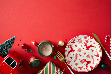 Festive table setting with plate in Christmas plate, cacao cup, truck with Christmas tree, baubles on red background. Top view.