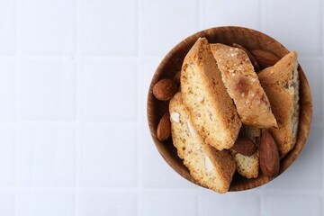 Tasty almond biscuits (Cantuccini) and nuts in bowl on white tiled table, top view. Space for text