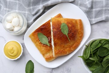 Pieces of tasty calzone with basil, mozzarella cheese and sauce on white marble table, flat lay