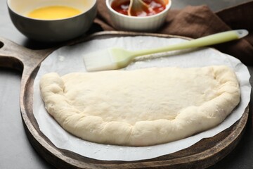 Uncooked calzone pizza, sauce and egg yolk on grey table, closeup