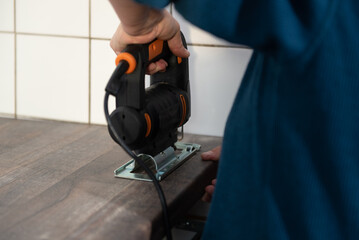 A woman is using a jigsaw to cut openings for electrical appliances into the kitchen countertop.