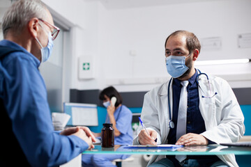 Young doctor consults with elderly male patient, attentively documenting symptoms on clipboard during medical checkup at clinic. Physician writing health concerns of retired man in hospital office.