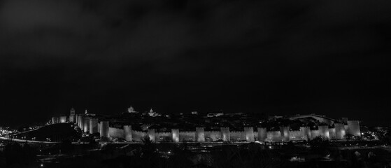 Illuminated nighttime scene showcasing the iconic medieval rampart wall and main entrance gate of &Aacute;vila, Spain. The architecture is beautifully highlighted by soft lighting surrounded by a quiet plaza