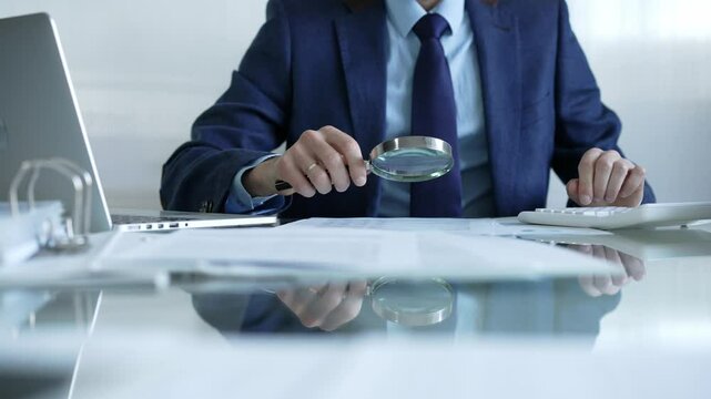 Businessman analyzing financial data with magnifying glass and calculator, conducting audit or review in office. Audit and taxes theme in business