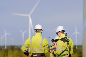 A team of engineers in high visibility safety gear stand together at a wind farm, conducting inspections and discussing renewable energy projects. Wind turbines surround them under a clear sky.