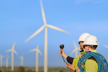 Two engineers wearing high visibility safety jackets and helmets measure wind speed with an anemometer and analyze data on a tablet at a renewable energy site. The cloudy sky adds depth to the scene.