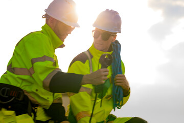 Two engineers wearing high visibility safety jackets and helmets measure wind speed with an anemometer and analyze data on a tablet at a renewable energy site. The cloudy sky adds depth to the scene.