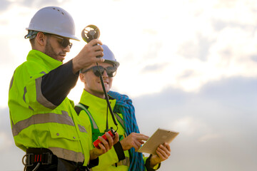 Two engineers wearing high visibility safety jackets and helmets measure wind speed with an...