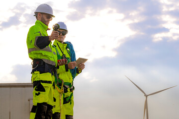 Two engineers wearing high visibility safety jackets and helmets measure wind speed with an anemometer and analyze data on a tablet at a renewable energy site. The cloudy sky adds depth to the scene.