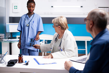 Black healthcare worker gives radiograph to older doctor as male pensioner patient waits in clinic...